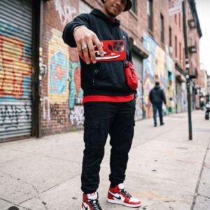 Young man in urban streetwear capturing selfie with smartphone against graffiti wall.